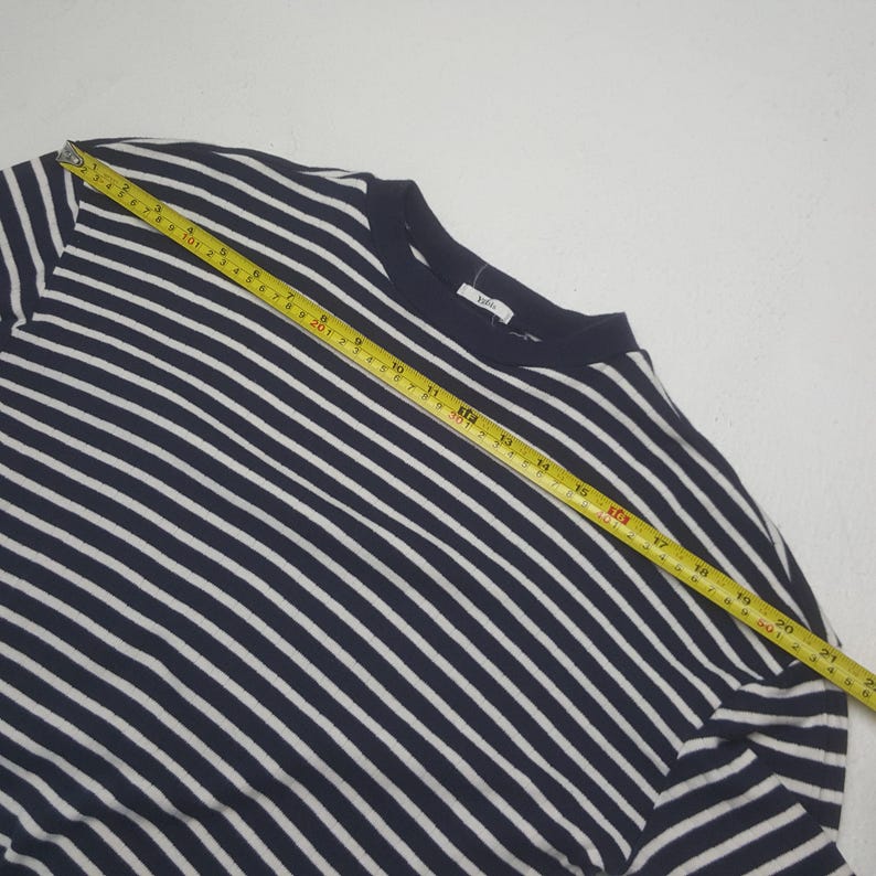 May include: Navy blue and white striped long-sleeve shirt. The shirt is measured with a yellow tape measure. The shirt has a crew neck and a tag on the back of the neck. The stripes are horizontal and evenly spaced.