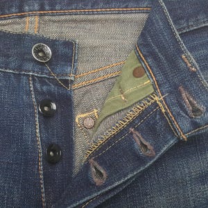May include: Close-up of blue denim jeans with button fly. The jeans have black buttons and copper rivets. The interior of the jeans is visible, showing a green fabric and the stitching details.