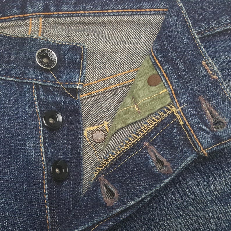 May include: Close-up of blue denim jeans with button fly. The jeans have black buttons and copper rivets. The interior of the jeans is visible, showing a green fabric and the stitching details.