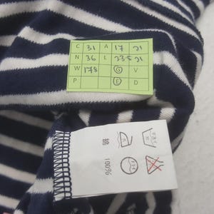 May include: Close-up of a navy blue and white striped fabric. A green tag with measurements and symbols is attached. A white tag with washing instructions is also visible. The fabric is 100% cotton.