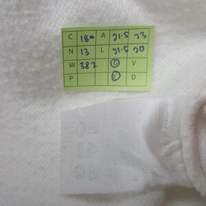 May include: A white fabric garment tag featuring a grid of laundry care symbols and a light green rectangular sticker with handwritten blue ink notes including C 18, N 13, W 382, A 21.5, L 21.5, and circled letters G and E on a textured white surface.