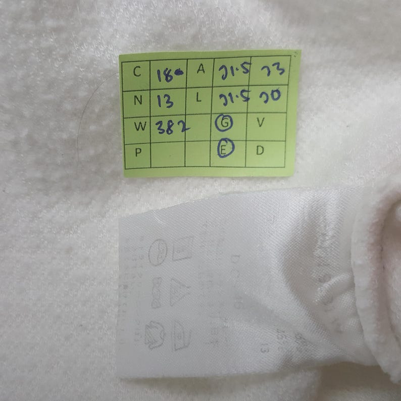 May include: A white fabric garment tag featuring a grid of laundry care symbols and a light green rectangular sticker with handwritten blue ink notes including C 18, N 13, W 382, A 21.5, L 21.5, and circled letters G and E on a textured white surface.