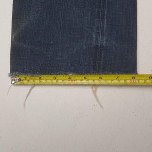 May include: Close-up of a section of dark blue denim fabric. A yellow measuring tape is laid across the bottom edge, showing measurements in inches. The denim has a frayed edge.