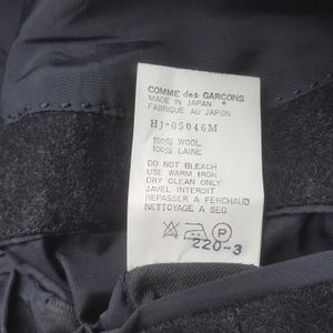 May include: A close-up of a tag from a Comme des Garcons garment. The tag is white with black text, including "MADE IN JAPAN" and care instructions. The fabric appears to be dark gray wool.