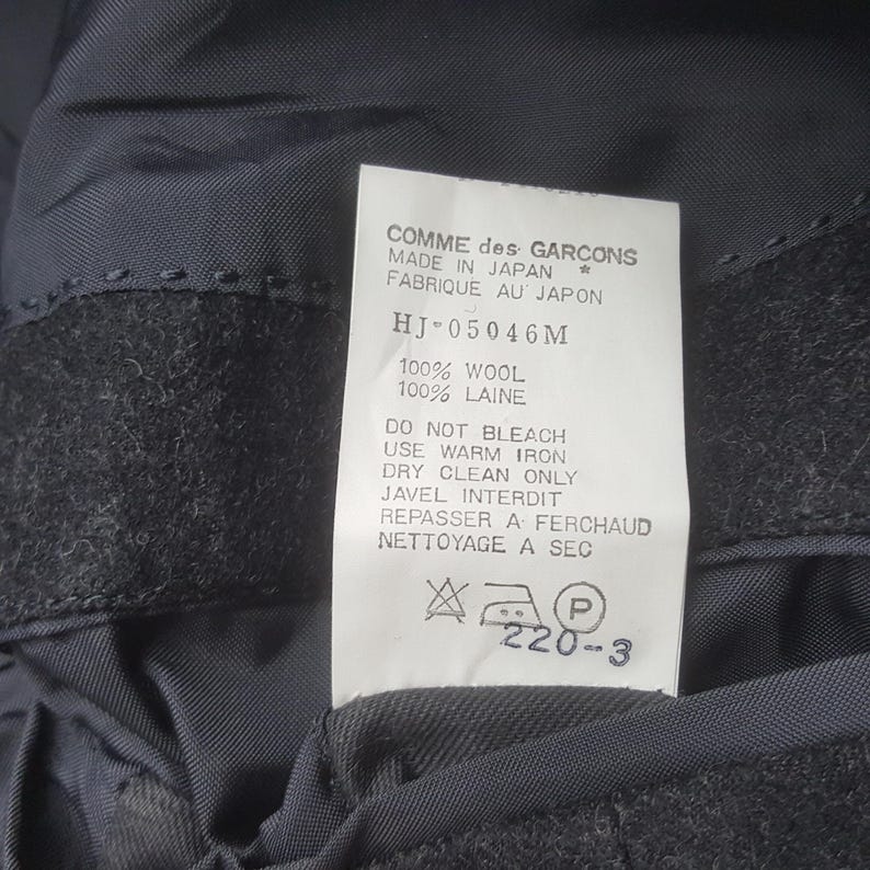 May include: A close-up of a tag from a Comme des Garcons garment. The tag is white with black text, including "MADE IN JAPAN" and care instructions. The fabric appears to be dark gray wool.