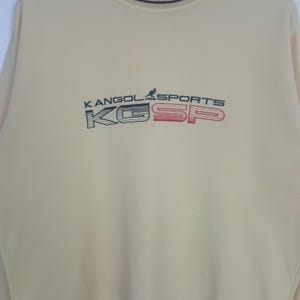 May include: A light beige sweatshirt with the text "KANGOL SPORTS" in navy blue and red. The sweatshirt has a crew neck and long sleeves. The letters "K" and "SP" are in a box design.