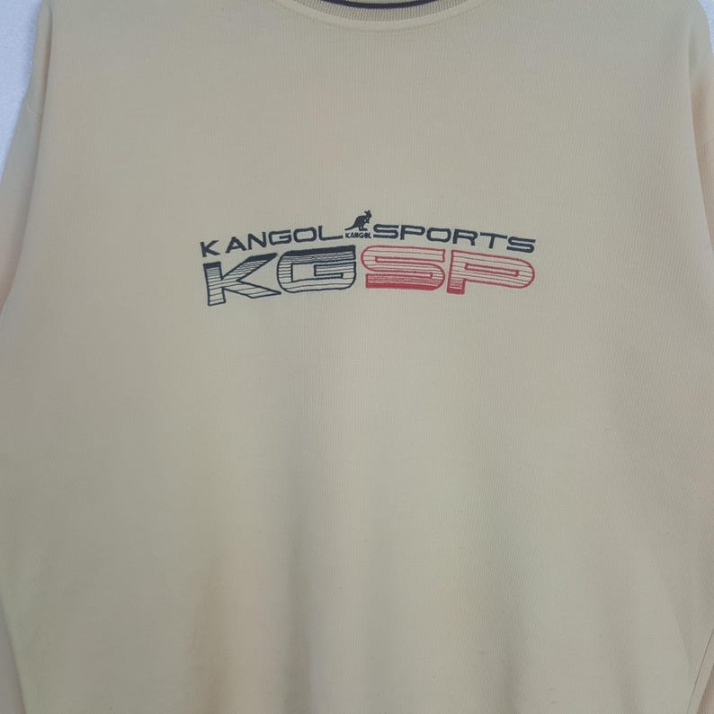 May include: A light beige sweatshirt with the text "KANGOL SPORTS" in navy blue and red. The sweatshirt has a crew neck and long sleeves. The letters "K" and "SP" are in a box design.