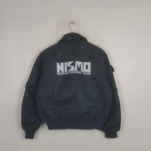 May include: A dark navy blue bomber jacket featuring the white text NISMO NISSAN RACING TEAM printed across the back. The jacket has a high collar, long sleeves with ribbed cuffs, and a ribbed hem, presented from a rear-facing perspective.