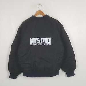 Puede incluir: Una chaqueta bomber negra con el texto blanco NISMO NISSAN RACING TEAM impreso en la espalda. La chaqueta tiene cuello, puños y dobladillo acanalados, confeccionada en una tela negra lisa y sólida con mangas largas.
