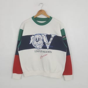 May include: White sweatshirt with a dog graphic and the text "STATUS MODERN Sports". The sweatshirt has a green collar, green and red accents on the sleeves, and a navy blue stripe across the chest.