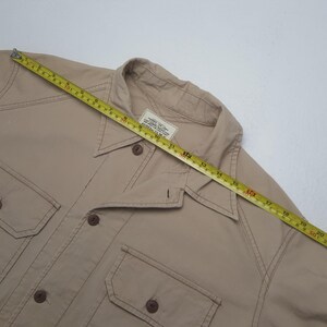 May include: A tan button-up shirt with two chest pockets and a collar. The shirt is being measured with a yellow measuring tape. The shirt has brown buttons and stitching. The shirt has a label on the inside.