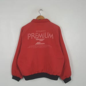 May include: A red jacket with a collar and long sleeves featuring black ribbed cuffs and a black hem. The back displays the white text MITSUBISHI PREMIUM Z with additional smaller printed text below the logo, all centered on the upper back panel.