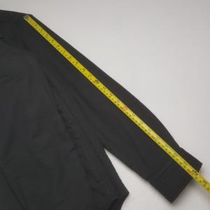 May include: A black long-sleeved shirt with a buttoned cuff is measured with a yellow tape measure. The tape measure shows measurements in inches. The shirt is made of a smooth fabric.