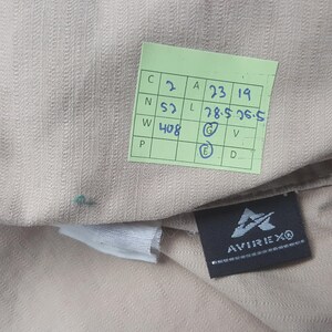 May include: Close-up of a tan fabric with a textured weave. A green tag with handwritten numbers and letters is visible, along with a black label that reads "AVIREXO" in white.