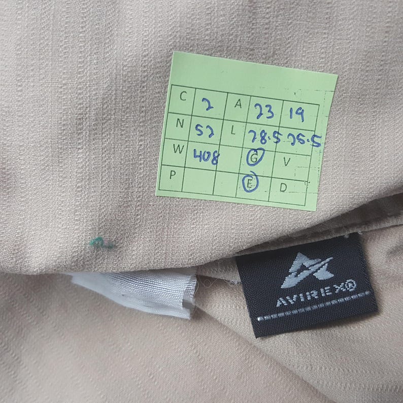 May include: Close-up of a tan fabric with a textured weave. A green tag with handwritten numbers and letters is visible, along with a black label that reads "AVIREXO" in white.