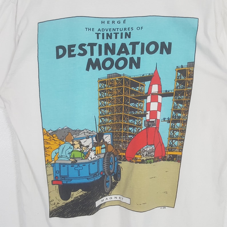 Puede incluir: Camiseta blanca con un gr&aacute;fico de "Las aventuras de Tint&iacute;n: Objetivo Luna". El dise&ntilde;o incluye un cohete rojo, un jeep azul y el texto del t&iacute;tulo. La camiseta es de cuello redondo.
