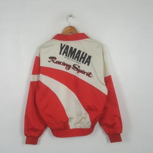 May include: A red and off-white long-sleeve jacket featuring the black text YAMAHA MOTOR CYCLE SPORTS and the red and black text Racing Spirit printed on the back. The jacket has a collar, ribbed cuffs, and a ribbed hem, hanging on a hanger.