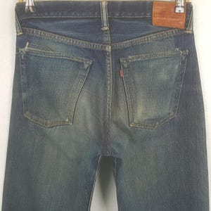 May include: Back view of blue denim jeans with two back pockets and a small red tag. The jeans have a worn, faded appearance with distressed details. A brown leather patch is visible at the waistband.