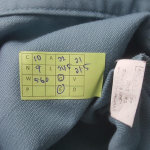K&ouml;nnte beinhalten: Ein blaues Kleidungsst&uuml;ck aus Stoff mit einem gelben rechteckigen Etikett mit handschriftlichen Ma&szlig;en in cm, darunter 10, 9, 560, 22, 24.5, 31 und 21.5. Ein wei&szlig;es Pflegeetikett mit japanischem Text und MADE IN THE PEOPLES ist seitlich angebracht.