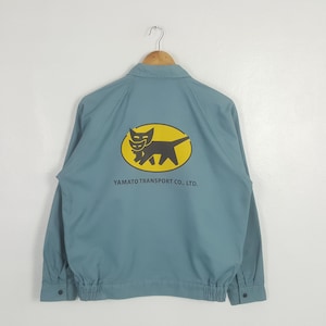 K&ouml;nnte beinhalten: Eine hellblaue Langarmjacke mit Kragen und elastischem Saum. Auf der R&uuml;ckseite befindet sich eine gelbe ovale Grafik mit der schwarzen Silhouette einer Katzenmutter, die ein K&auml;tzchen tr&auml;gt, sowie der Aufschrift YAMATO TRANSPORT CO., LTD.