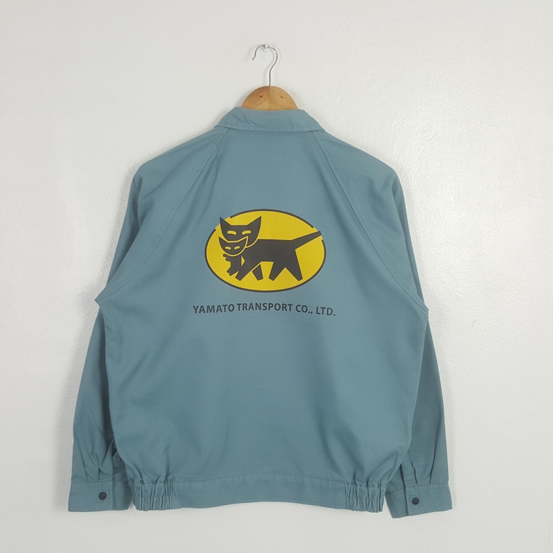 K&ouml;nnte beinhalten: Eine hellblaue Langarmjacke mit Kragen und elastischem Saum. Auf der R&uuml;ckseite befindet sich eine gelbe ovale Grafik mit der schwarzen Silhouette einer Katzenmutter, die ein K&auml;tzchen tr&auml;gt, sowie der Aufschrift YAMATO TRANSPORT CO., LTD.