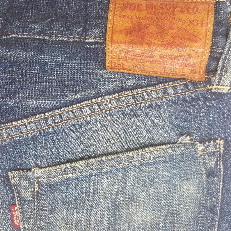 May include: Close-up of blue denim jeans with a brown leather patch. The patch reads "JOE McCOY & CO. REAL WORK CLOTHING XH" and includes text "Lot 901 29 L 36". The jeans have a back pocket and visible stitching.