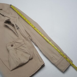 May include: Tan jacket with visible stitching and a buttoned cuff. A yellow measuring tape is laid across the jacket, showing measurements in inches. The jacket has a small embroidered design on the sleeve.