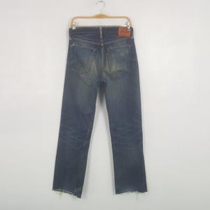 May include: Dark wash denim jeans with a distressed look, featuring a raw hem. The jeans are displayed on a hanger, showcasing the back pockets and a leather patch. The denim has a faded appearance.