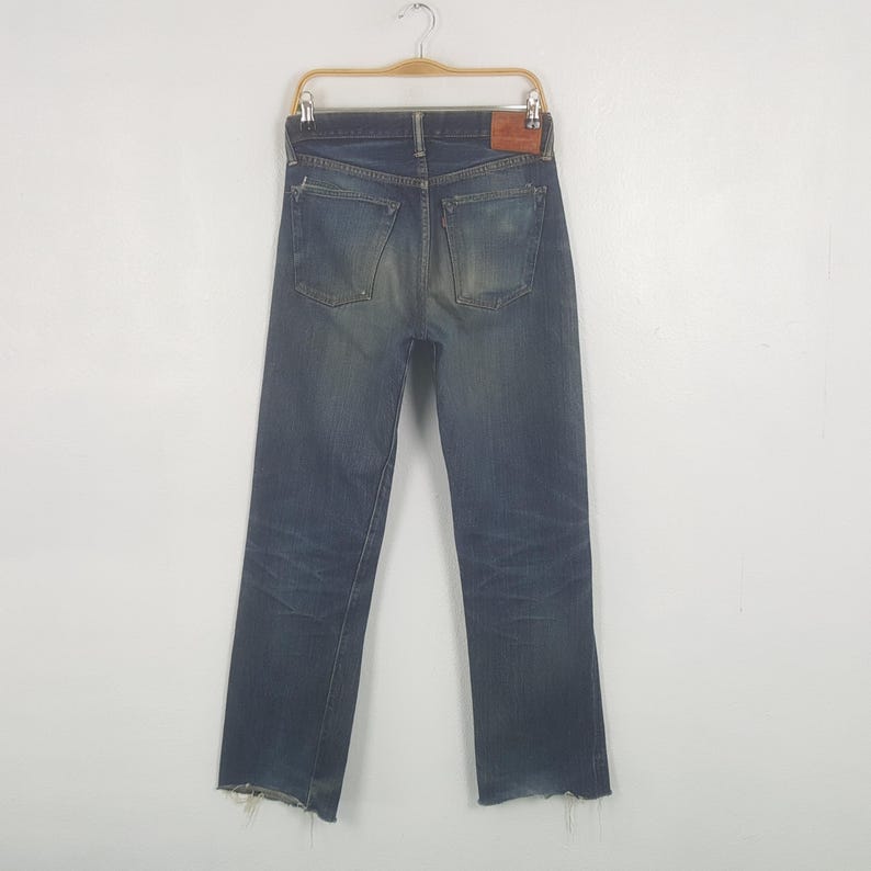 May include: Dark wash denim jeans with a distressed look, featuring a raw hem. The jeans are displayed on a hanger, showcasing the back pockets and a leather patch. The denim has a faded appearance.