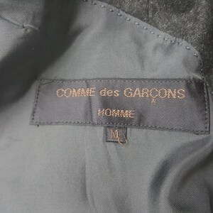 May include: Close-up of a dark gray garment's interior, showcasing a label with the text "COMME des GAR&Ccedil;ONS HOMME" in gold lettering. Below the text is a size "M" tag.