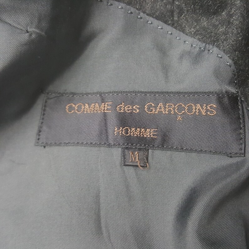 May include: Close-up of a dark gray garment's interior, showcasing a label with the text "COMME des GAR&Ccedil;ONS HOMME" in gold lettering. Below the text is a size "M" tag.