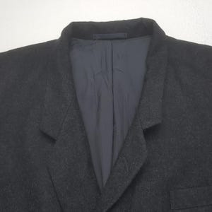 May include: A dark gray blazer with a notched lapel and a single-button closure. The interior lining is a dark blue fabric. The blazer is made of a textured material, and the image is a close-up of the upper portion of the garment.