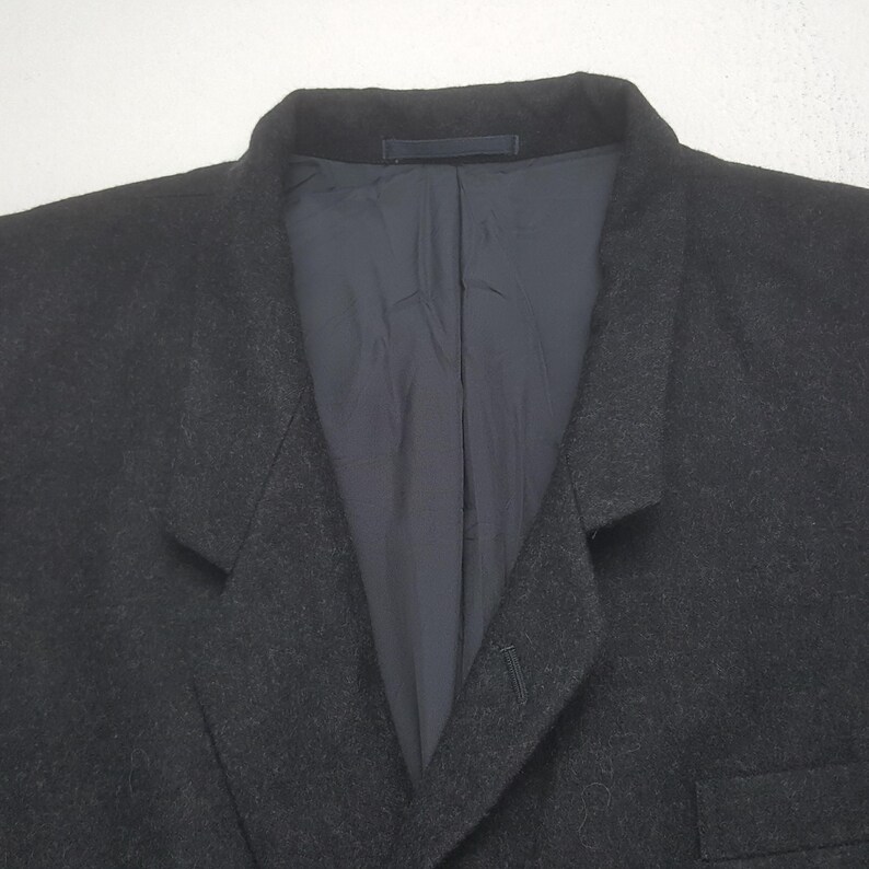 May include: A dark gray blazer with a notched lapel and a single-button closure. The interior lining is a dark blue fabric. The blazer is made of a textured material, and the image is a close-up of the upper portion of the garment.