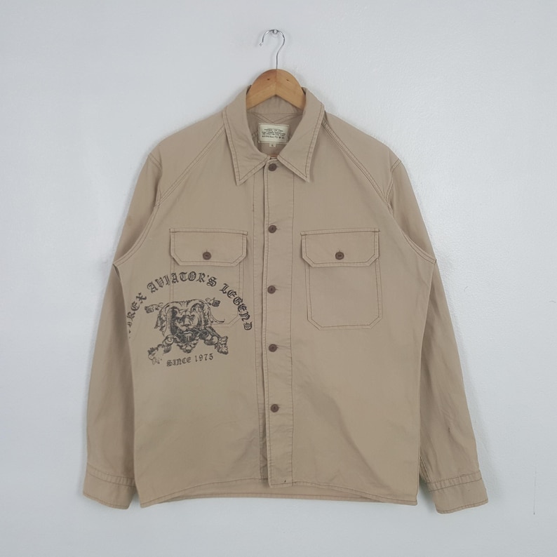 May include: Tan button-up shirt with two chest pockets and a collar. The shirt features a graphic on the left side that reads "Rex Aviator's Legend" with an illustration and "since 1975". The shirt is hanging on a wooden hanger.