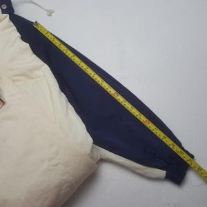 Pode incluir: Casaco creme e azul marinho com capuz e cord&atilde;o. A manga &eacute; medida com uma fita m&eacute;trica amarela, mostrando um comprimento de aproximadamente 56 cm.