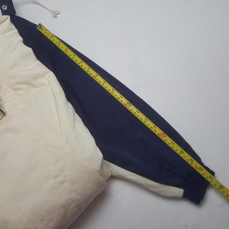 Pode incluir: Casaco creme e azul marinho com capuz e cord&atilde;o. A manga &eacute; medida com uma fita m&eacute;trica amarela, mostrando um comprimento de aproximadamente 56 cm.