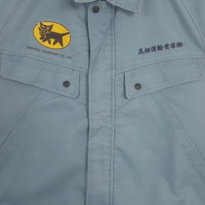 K&ouml;nnte beinhalten: Ein hellblaues Arbeitshemd mit einem gelben ovalen Logo, das eine schwarze Katzengrafik und den Text YAMATO TRANSPORT CO., LTD. auf der linken Brust zeigt. Auf der rechten Brust befindet sich japanischer Text in Schwarz. Zwei gekn&ouml;pfte Taschen.
