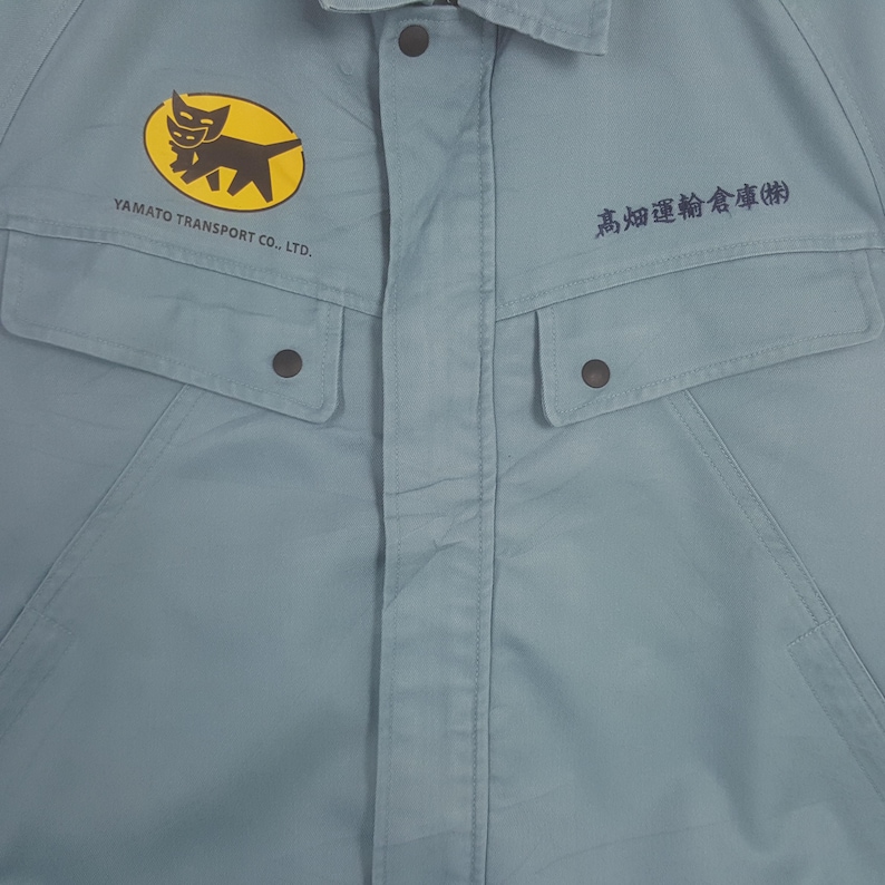 K&ouml;nnte beinhalten: Ein hellblaues Arbeitshemd mit einem gelben ovalen Logo, das eine schwarze Katzengrafik und den Text YAMATO TRANSPORT CO., LTD. auf der linken Brust zeigt. Auf der rechten Brust befindet sich japanischer Text in Schwarz. Zwei gekn&ouml;pfte Taschen.