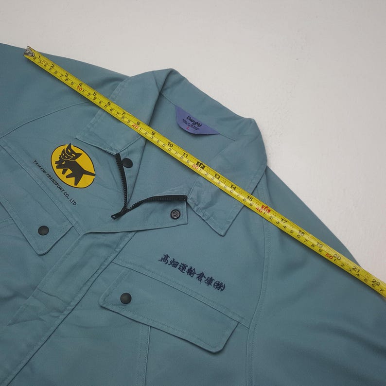 K&ouml;nnte beinhalten: Eine hellblaue Arbeitsjacke mit einem gelben kreisf&ouml;rmigen Logo, das eine schwarze Katze und den Text TAMAKI TRANSPORT CO., LTD. zeigt. Ein gelbes Ma&szlig;band liegt &uuml;ber der Brust und zeigt eine Breite von etwa 56 cm. Schwarzer Text ist aufgedruckt.