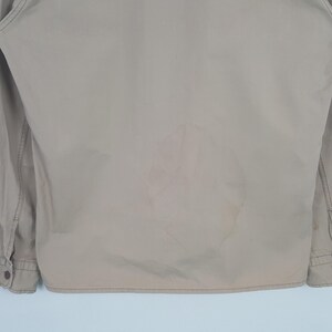 May include: Tan-colored jacket with long sleeves and buttoned cuffs. The back of the jacket has a large, lighter-colored stain. The jacket is made of a smooth fabric and appears to be lightly worn.