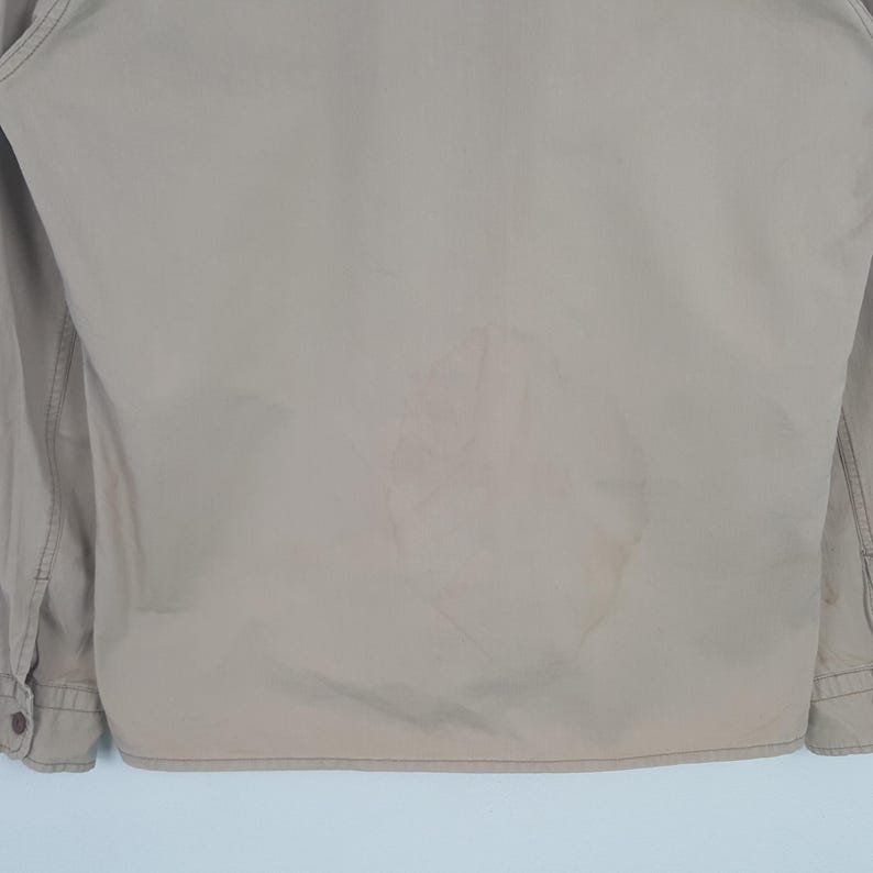 May include: Tan-colored jacket with long sleeves and buttoned cuffs. The back of the jacket has a large, lighter-colored stain. The jacket is made of a smooth fabric and appears to be lightly worn.