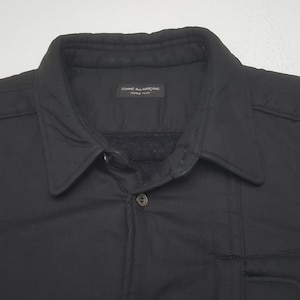 May include: Black button-down jacket with a collar. The jacket has a quilted design and a small pocket. The label reads "COMME des GARCONS HOMME PLUS".