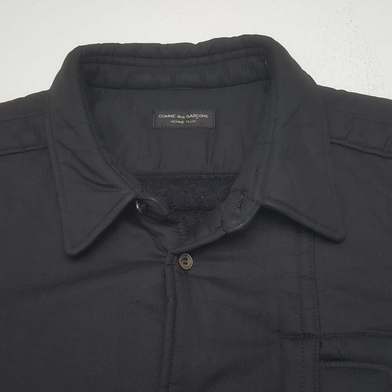 May include: Black button-down jacket with a collar. The jacket has a quilted design and a small pocket. The label reads "COMME des GARCONS HOMME PLUS".