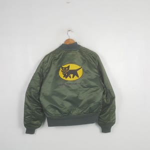 Puede incluir: Una chaqueta bomber de color verde oliva con un parche ovalado amarillo en la espalda que muestra un gráfico negro de dos gatos. El texto YAMATO TRANSPORT CO. LTD. está impreso en negro debajo del gráfico. La chaqueta tiene cuello, puños y dobladillo acanalados.