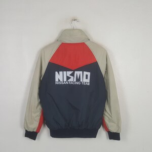 May include: A beige, red, and navy blue jacket featuring a high collar and long sleeves. The back displays the white text NISMO NISSAN RACING TEAM printed across a navy blue panel, with a red triangular accent above and beige shoulders.