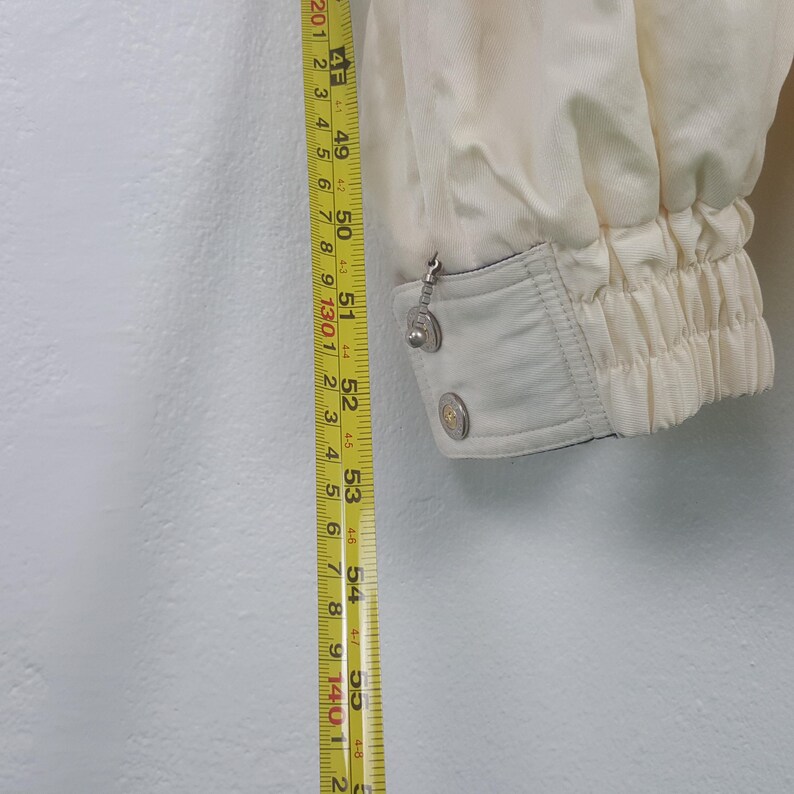 Pode incluir: Manga de casaco creme com punho el&aacute;stico e bot&otilde;es prateados. Uma fita m&eacute;trica mostra o comprimento do casaco. O casaco &eacute; bege claro e tem um aspeto texturizado. A manga tem aproximadamente 135 cm de comprimento.