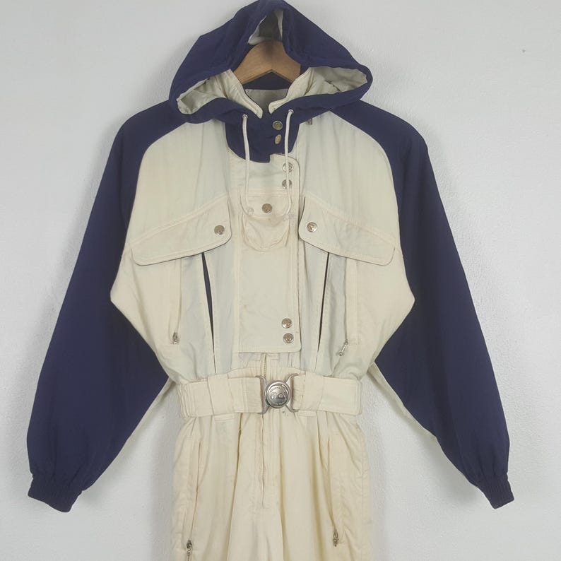 Pode incluir: Um fato de esqui vintage em creme e azul marinho. O fato de uma pe&ccedil;a apresenta um top com capuz com frente creme e mangas azul marinho. Possui v&aacute;rios bolsos, um cinto com fivela prateada e fecho de correr. Uma pe&ccedil;a de roupa de inverno cl&aacute;ssica.
