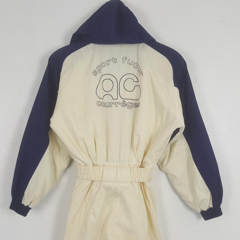 Pode incluir: Macac&atilde;o vintage creme com mangas e capuz azul marinho. A parte de tr&aacute;s apresenta o texto "sport futur" acima do logotipo "AC Courr&egrave;ges" em preto. O macac&atilde;o tem cintura ajustada e design de corpo inteiro.