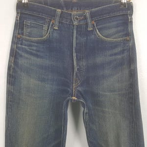 May include: Close-up of blue denim jeans with a faded wash. The jeans have a classic five-pocket design, a button fly, and belt loops. The denim shows signs of wear and fading, giving them a vintage look. The jeans are hanging on a hanger.