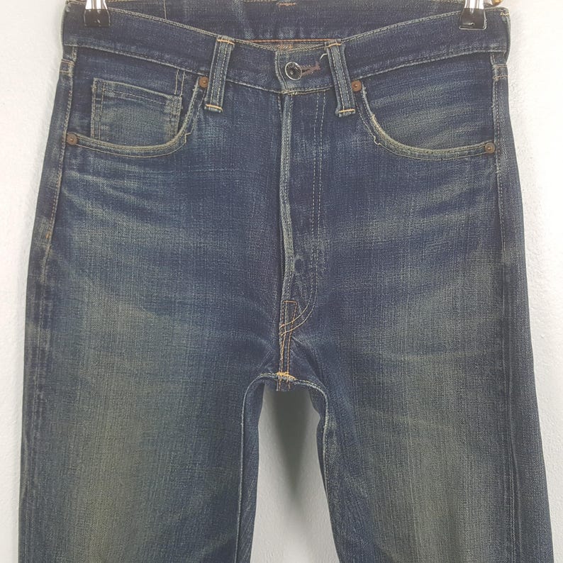 May include: Close-up of blue denim jeans with a faded wash. The jeans have a classic five-pocket design, a button fly, and belt loops. The denim shows signs of wear and fading, giving them a vintage look. The jeans are hanging on a hanger.